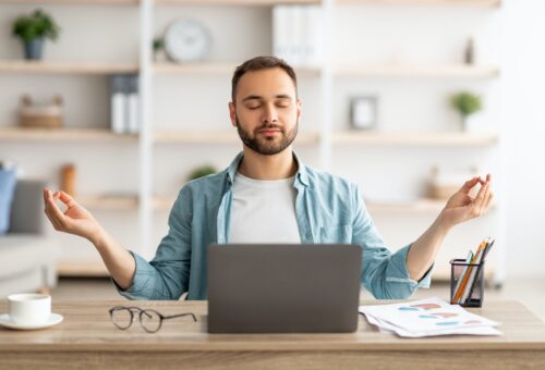 Workplace stress management. Calm Caucasian man meditating with closed eyes in front of laptop pc at