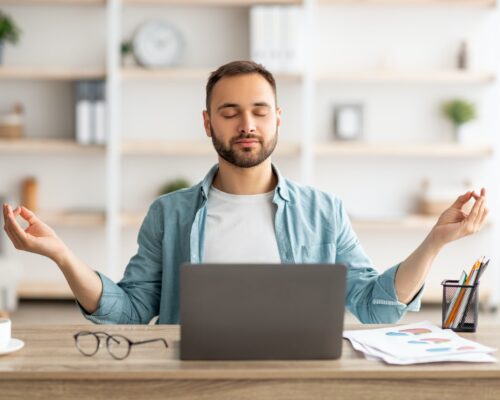 Workplace stress management. Calm Caucasian man meditating with closed eyes in front of laptop pc at